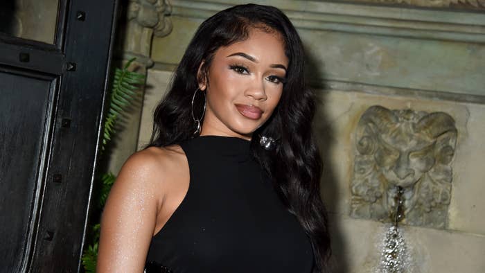 Saweetie at an event, wearing an elegant sleeveless black dress with long wavy hair, standing near a decorative stone wall