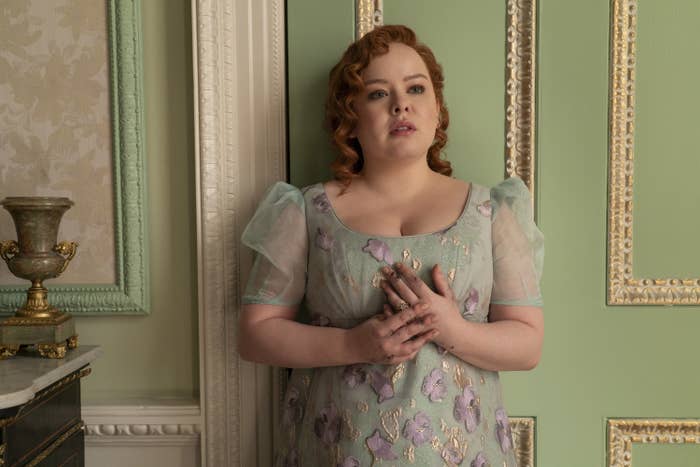 Nicola Coughlan as Penelope stands against a wall, wearing a vintage-style dress with floral patterns, looking contemplative with hands clasped