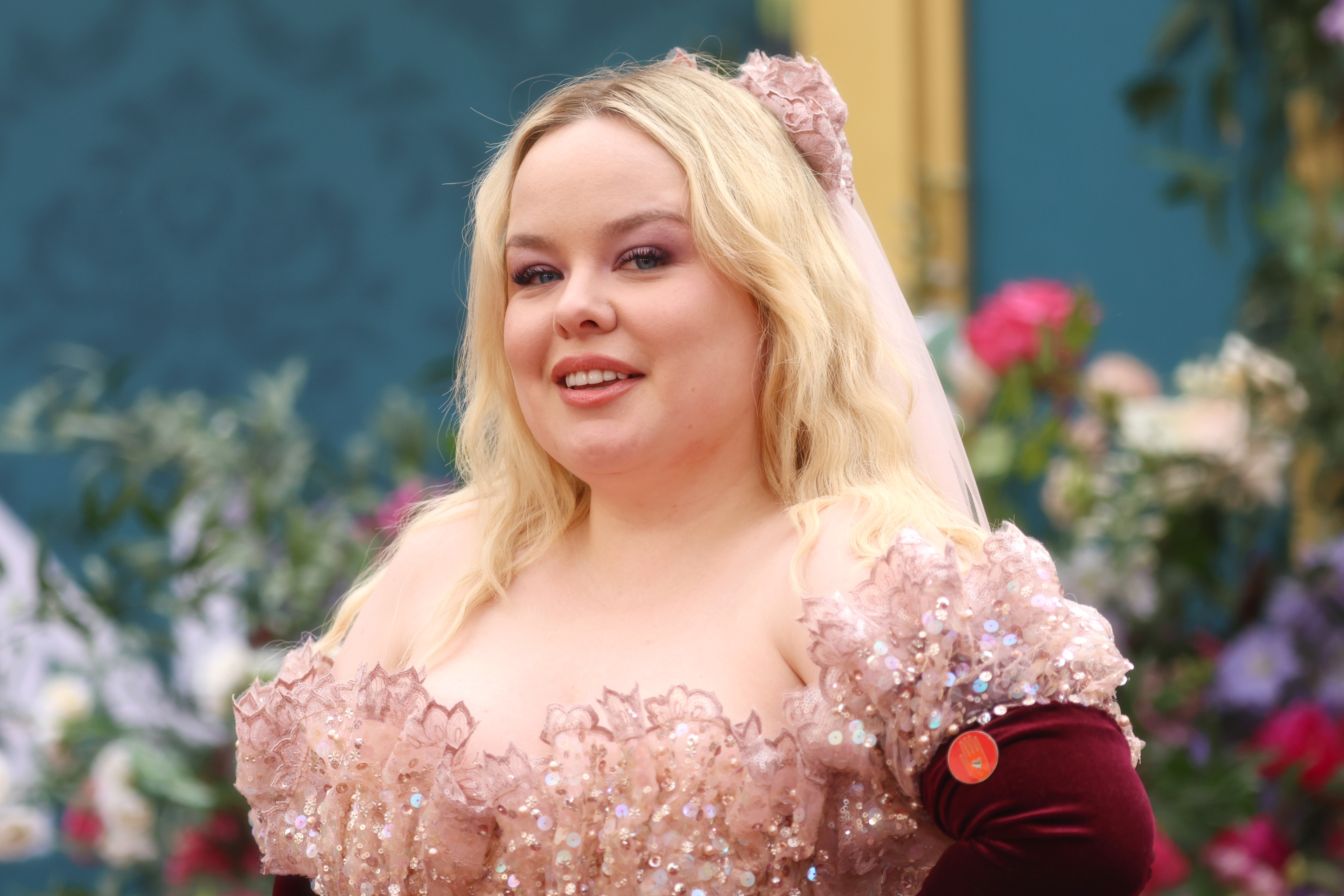 Nicola Coughlan is wearing an off-shoulder, sequined gown with floral details at a formal event, posing in front of a floral backdrop