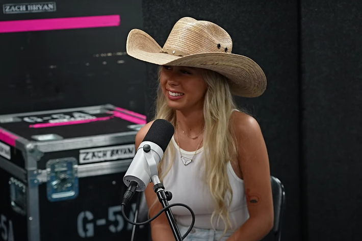 Person with long hair wearing a cowboy hat and sleeveless top sits at a microphone in a recording studio. Name on equipment reads "Zach Bryan."
