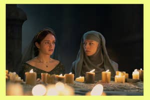 Olivia Cooke in a dark dress and headpiece, and Emma D'arcy in a gray hooded outfit, sit among numerous lit candles