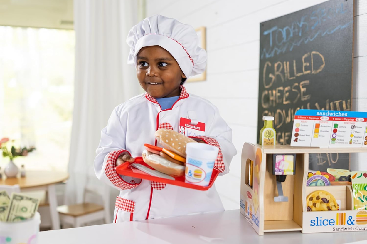 A young child dressed as a chef holds a toy sandwich, smoothie, and a tray at a play kitchen setup with a toy register
