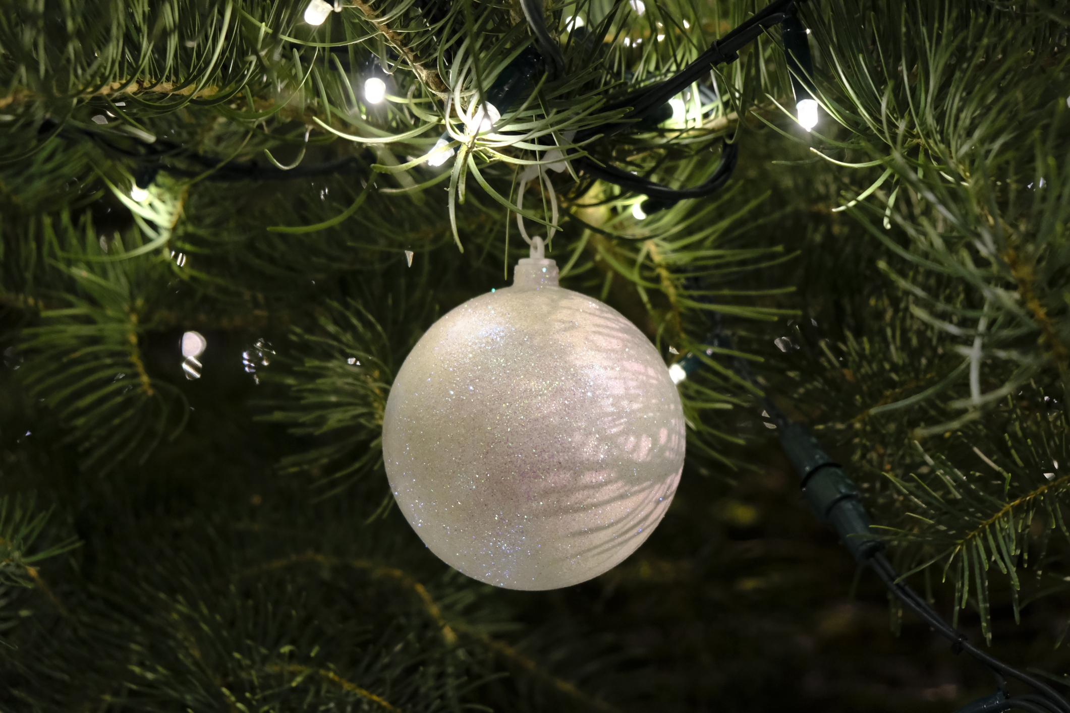 A close-up of a Christmas tree ornament hanging on a branch. The ornament is a shiny, textured ball. The background shows tree lights and green pine needles