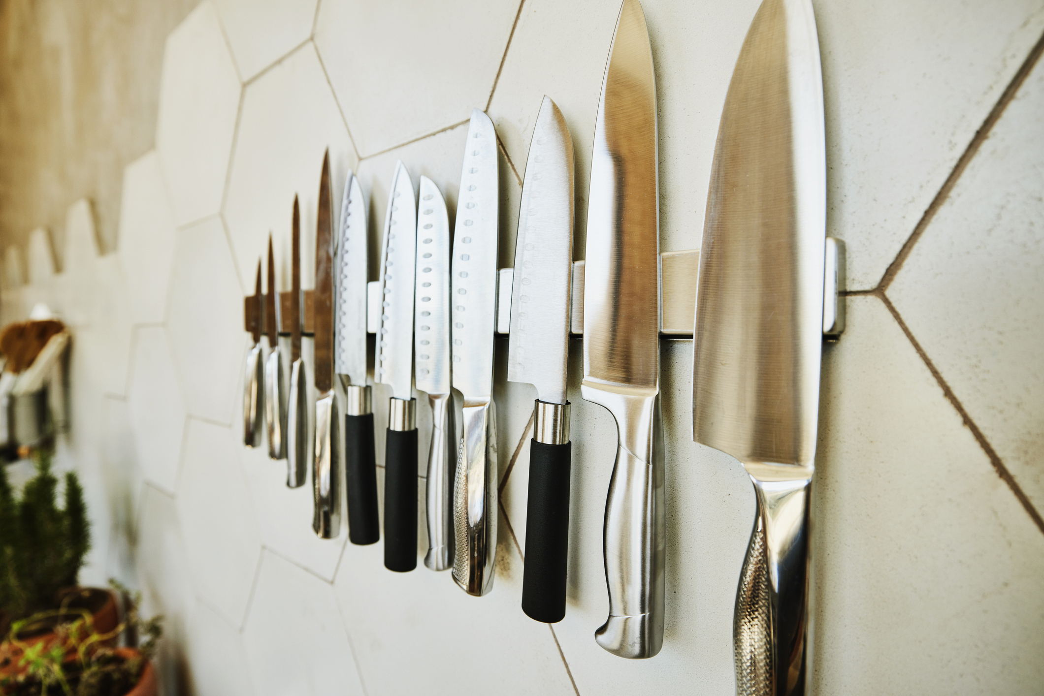 A row of kitchen knives neatly arranged on a magnetic strip mounted on a tiled kitchen wall. Various knife sizes and handles are visible