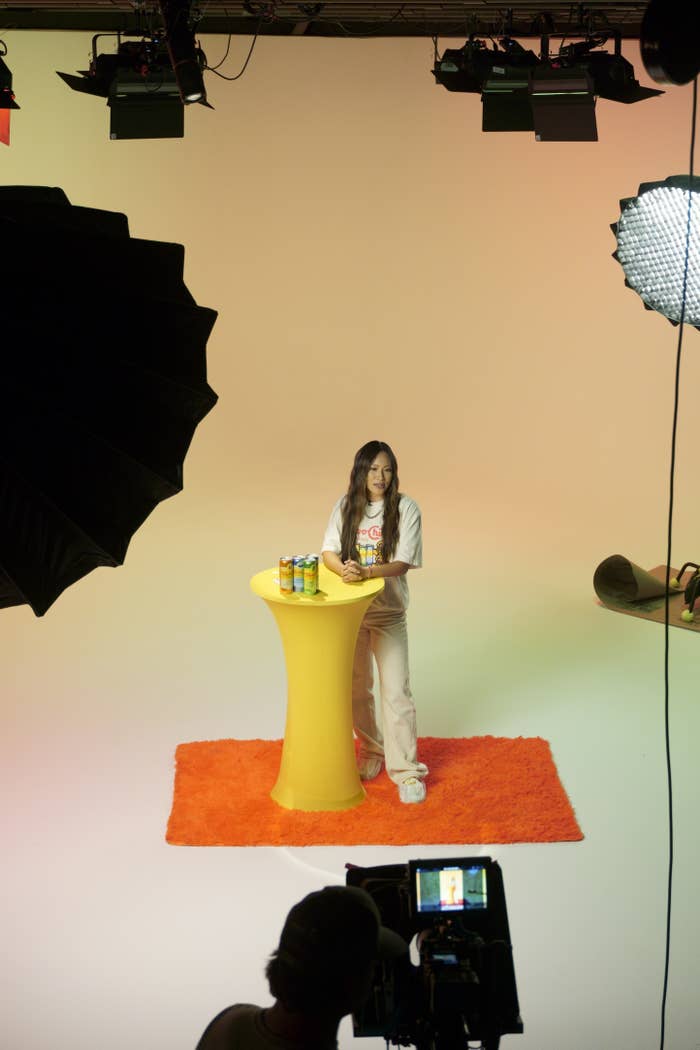 Olivia Rodrigo stands behind a tall yellow table with drinks on it, on an orange rug, during a photo shoot. She is wearing casual clothing
