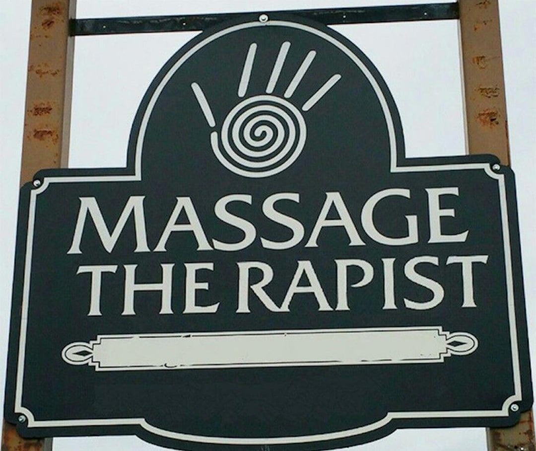 An outdoor sign that reads &quot;Massage the rapist&quot; with a decorative hand symbol at the top; there's some very unfortunate spacing going on (it's supposed to read Massage Therapist)