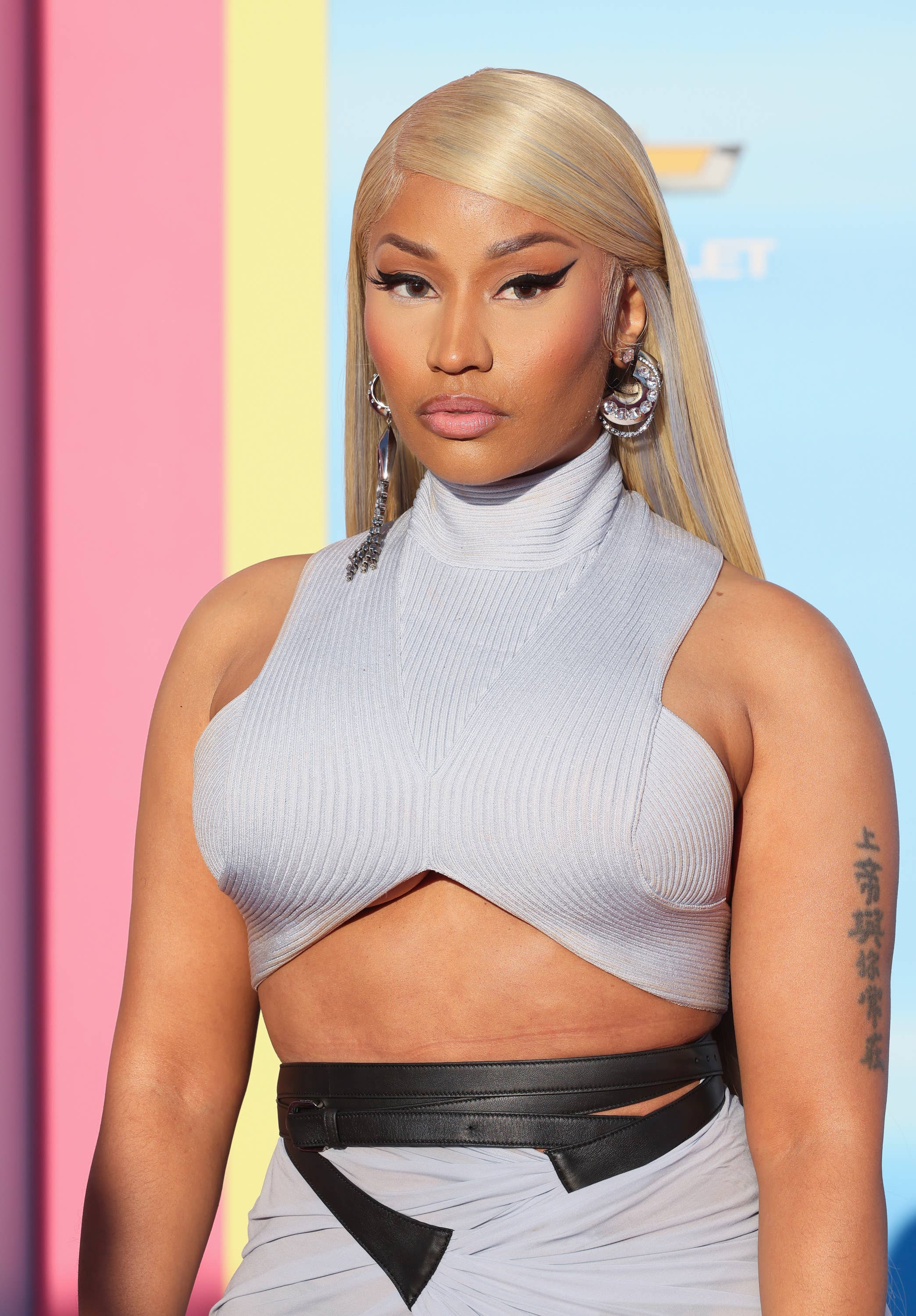 Nicki Minaj on the red carpet wearing a sleeveless high-neck crop top and high-waisted skirt with cut-out accents, accessorized with large hoop earrings