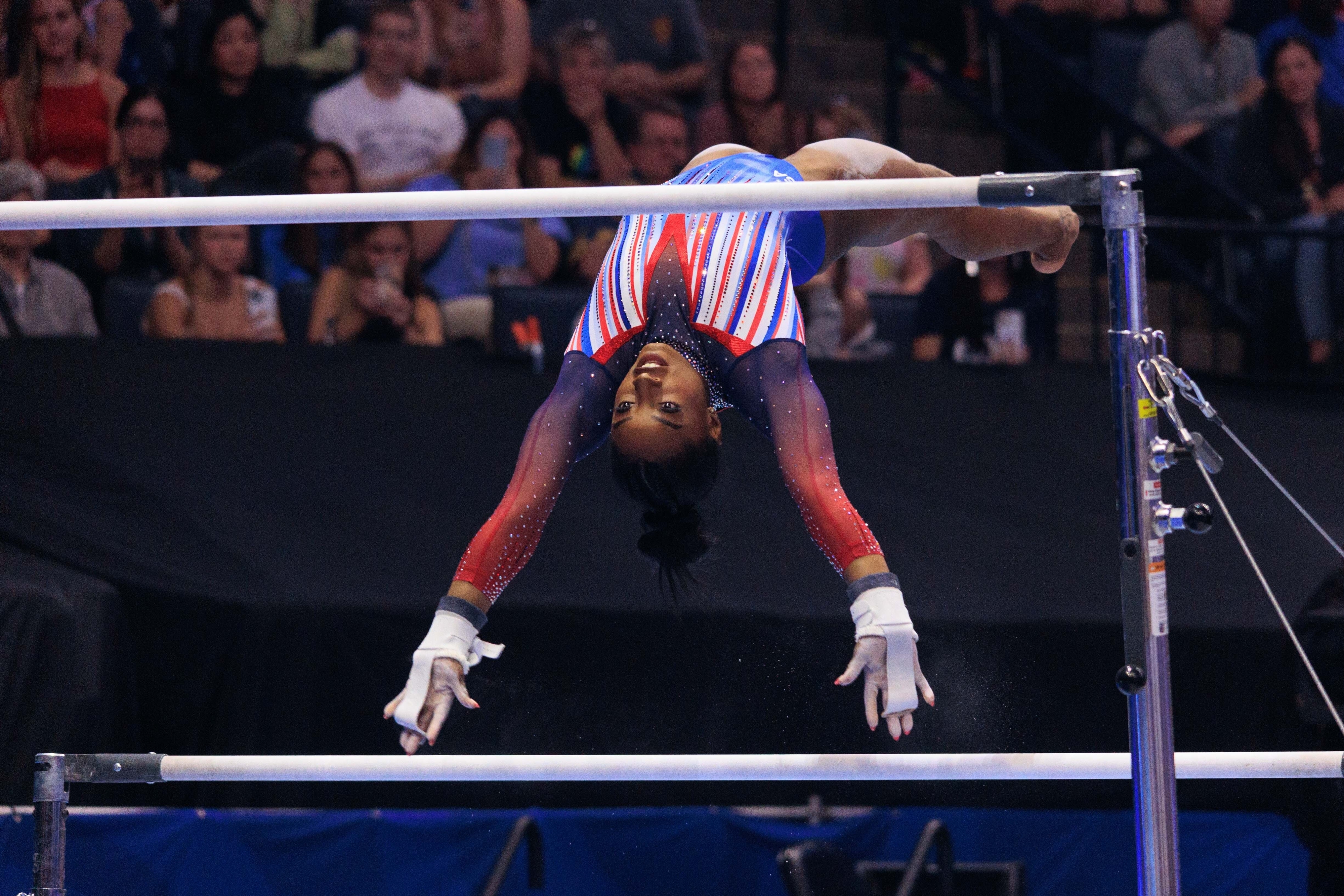 Simone Biles performs a complex routine on the uneven bars during a gymnastics competition, with an audience watching in the background