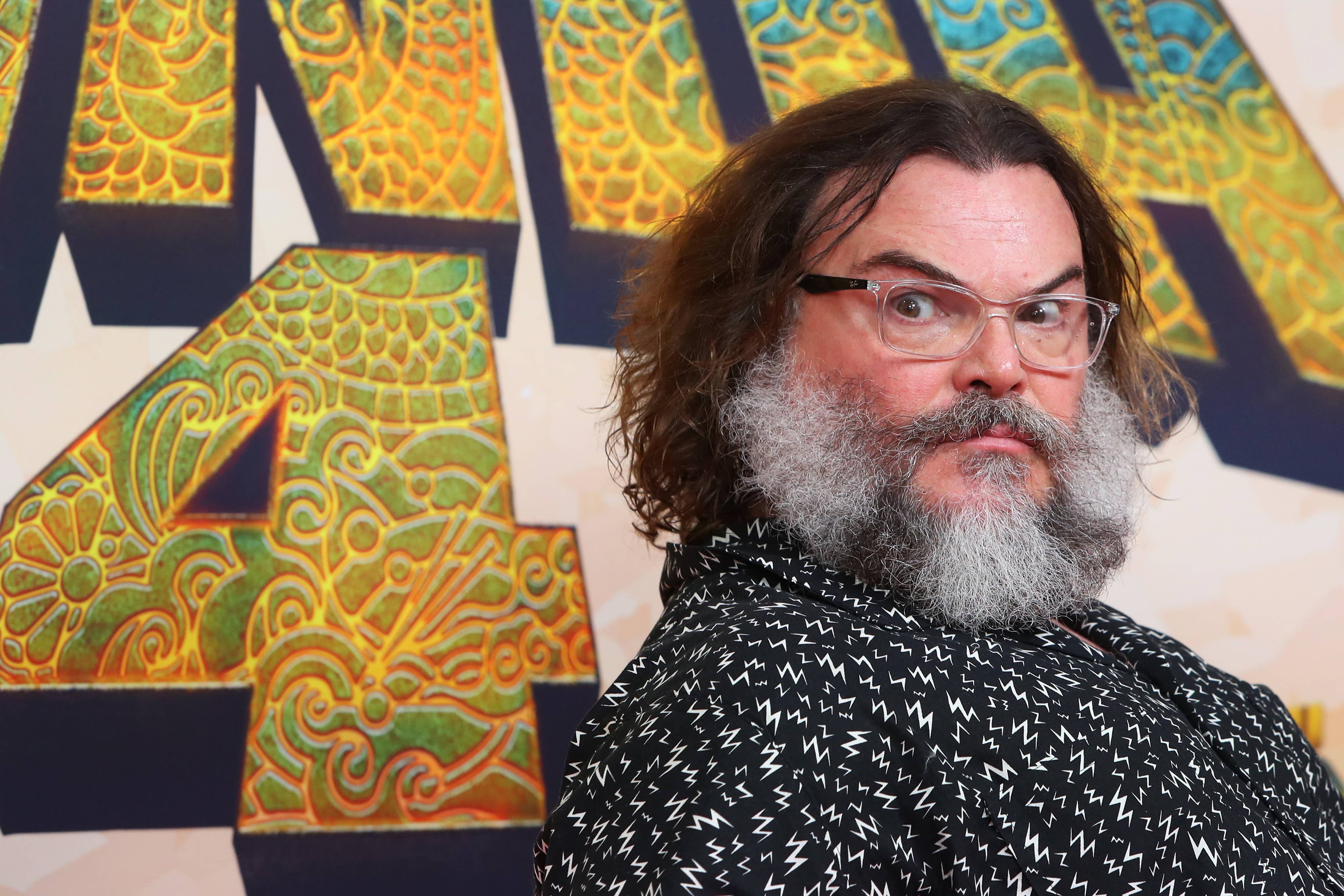 Jack Black poses on the red carpet, wearing a black patterned shirt, in front of an ornate background at a public event