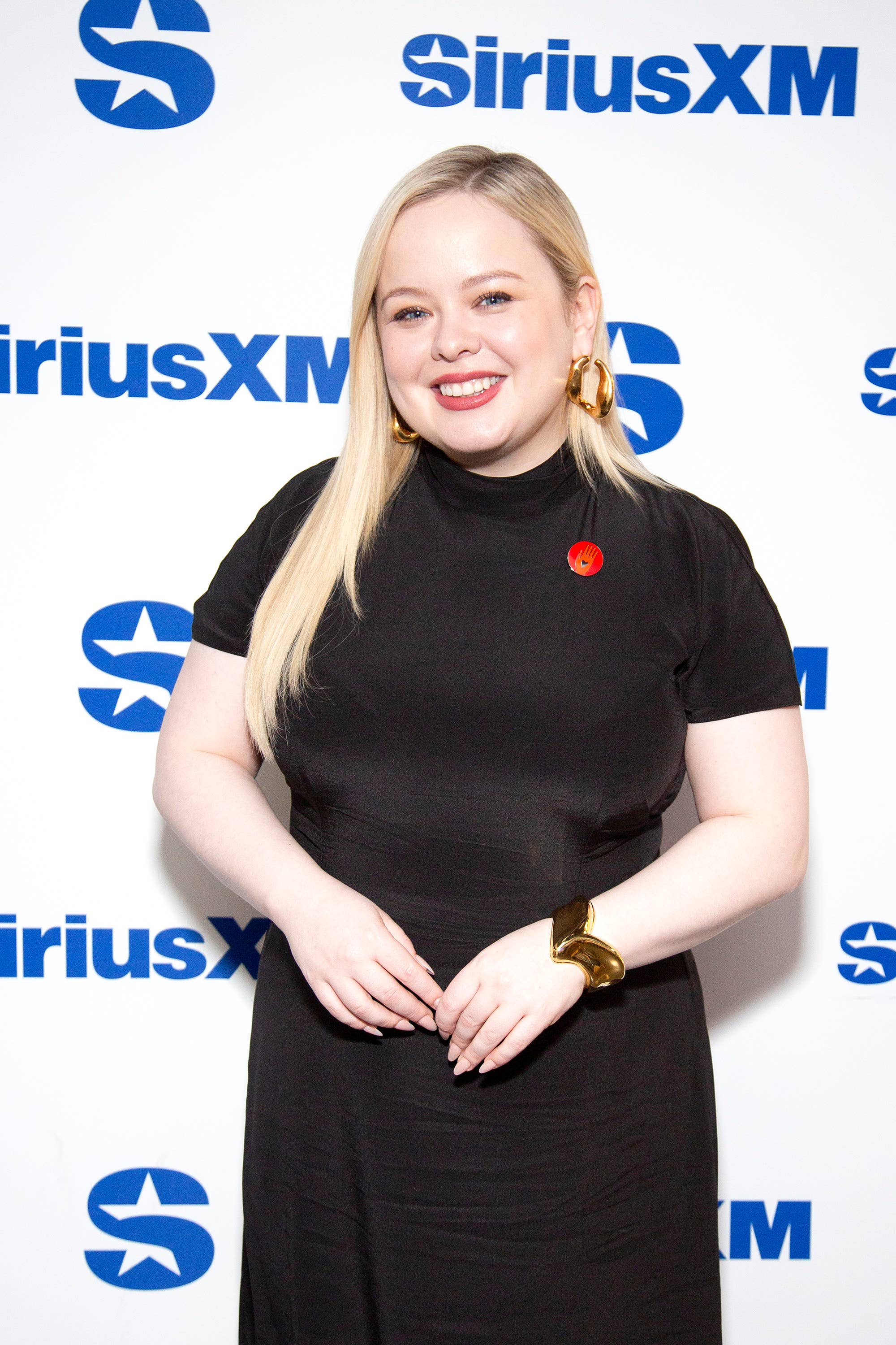 Nicola Coughlan smiles in a black dress with gold earrings and bracelet, posing in front of a SiriusXM backdrop