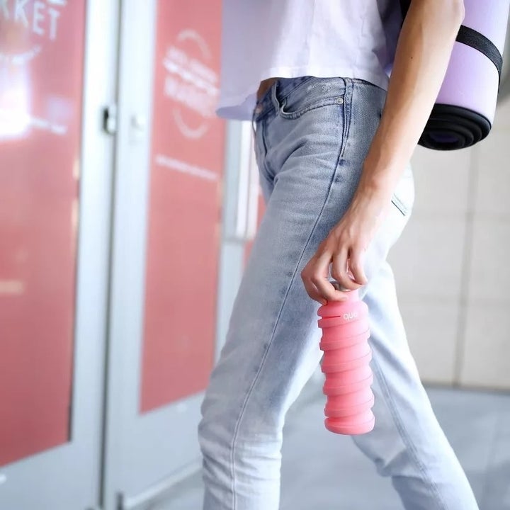 Person wearing jeans and a white shirt holding a pink collapsible water bottle with a yoga mat on their shoulder