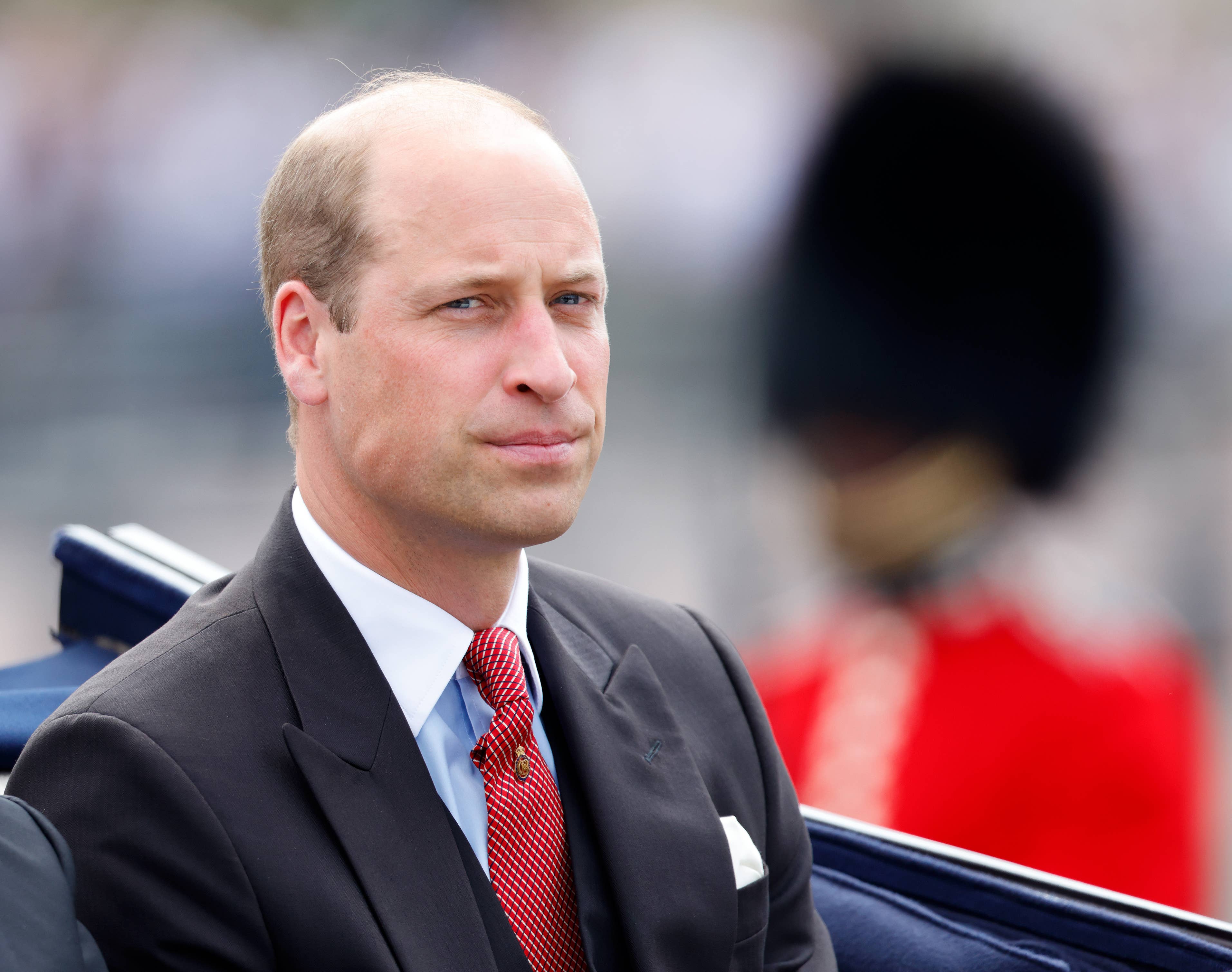 Prince William in a suit with a red tie attending an official event, a royal guard in full uniform is in the background