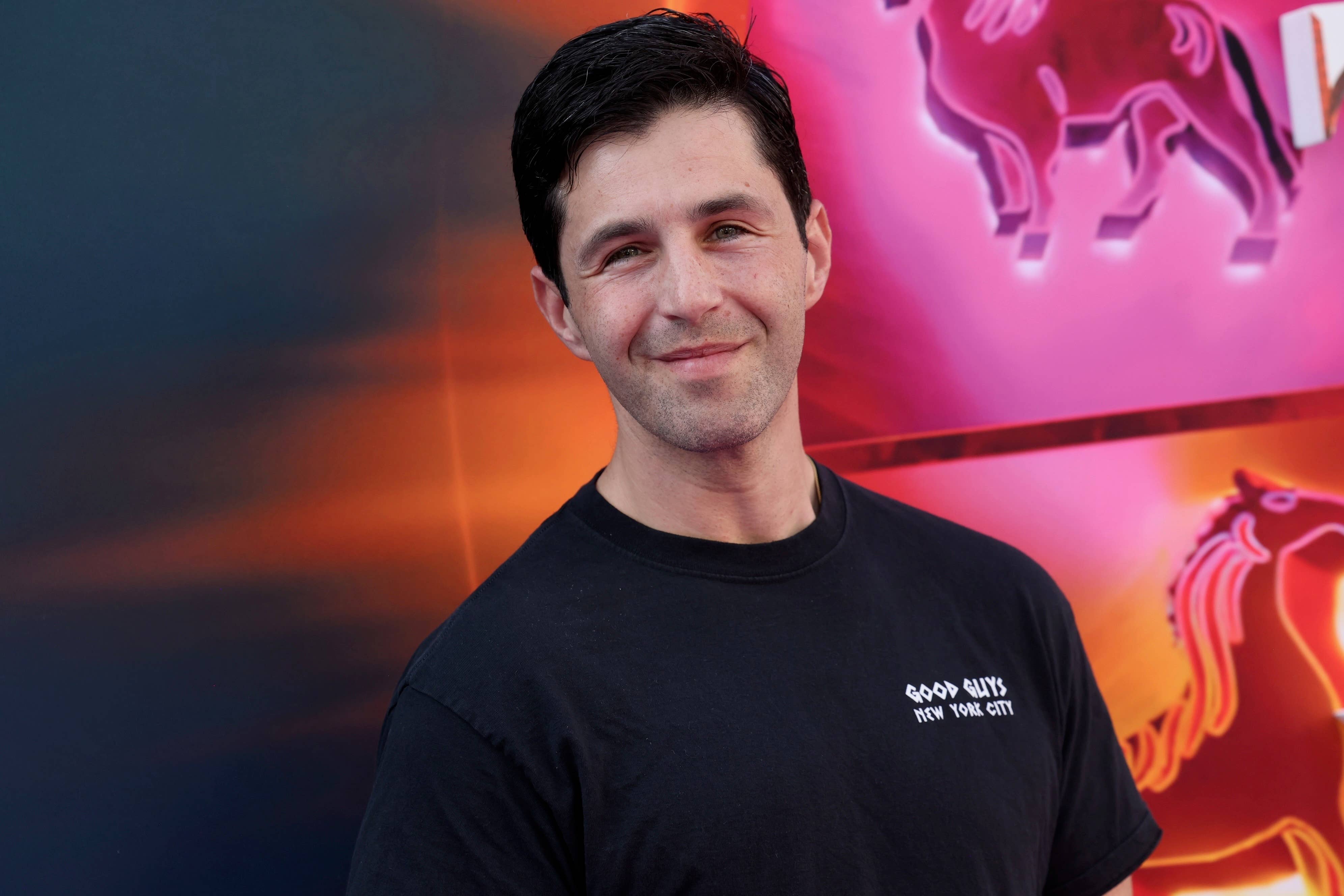 A person at a public event wearing a black t-shirt that reads "Good Guys New York City" is standing in front of a colorful backdrop featuring neon-lit horse images