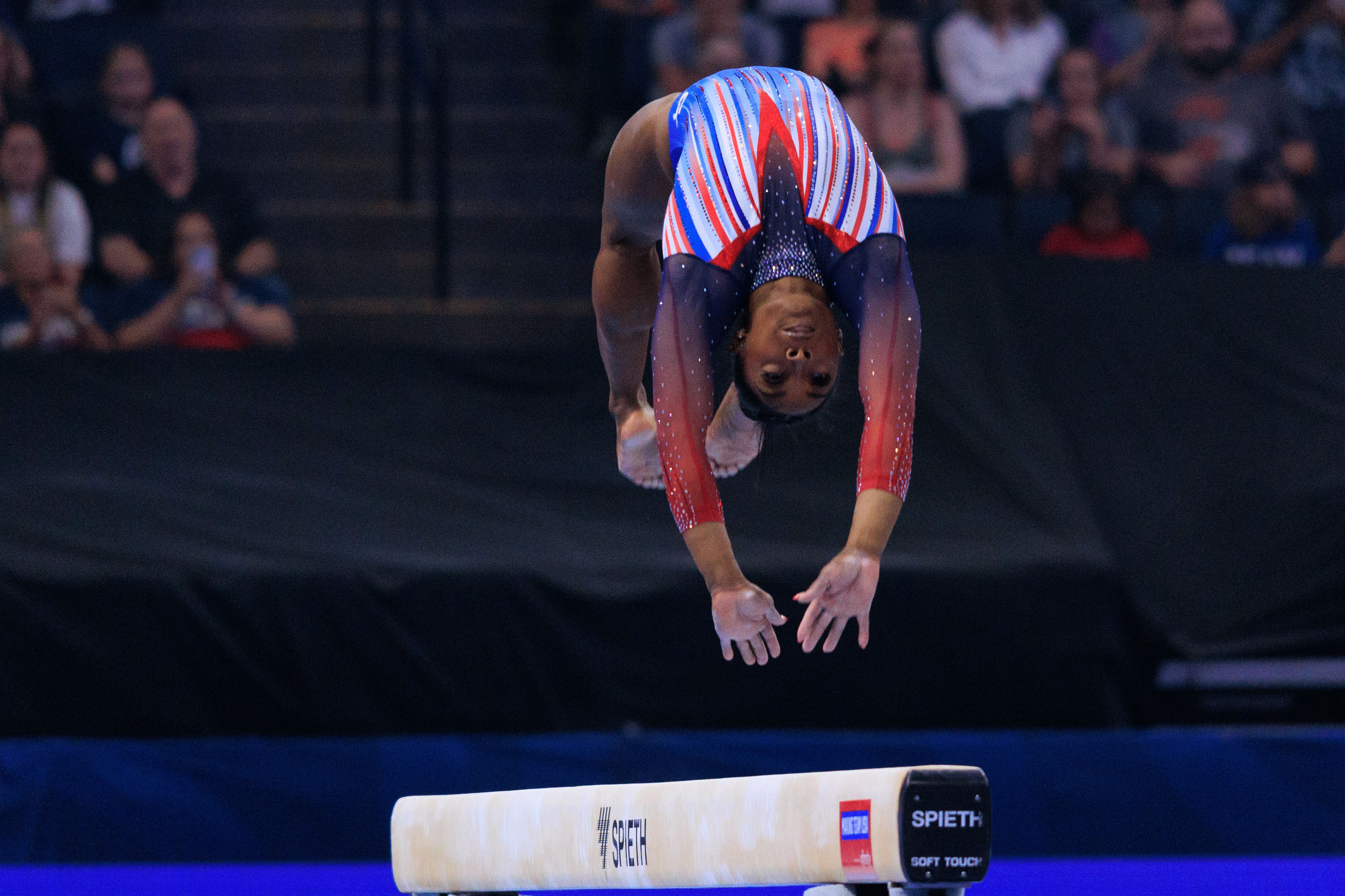 Simone Biles performs a backflip on the balance beam at a gymnastics competition with an audience in the background