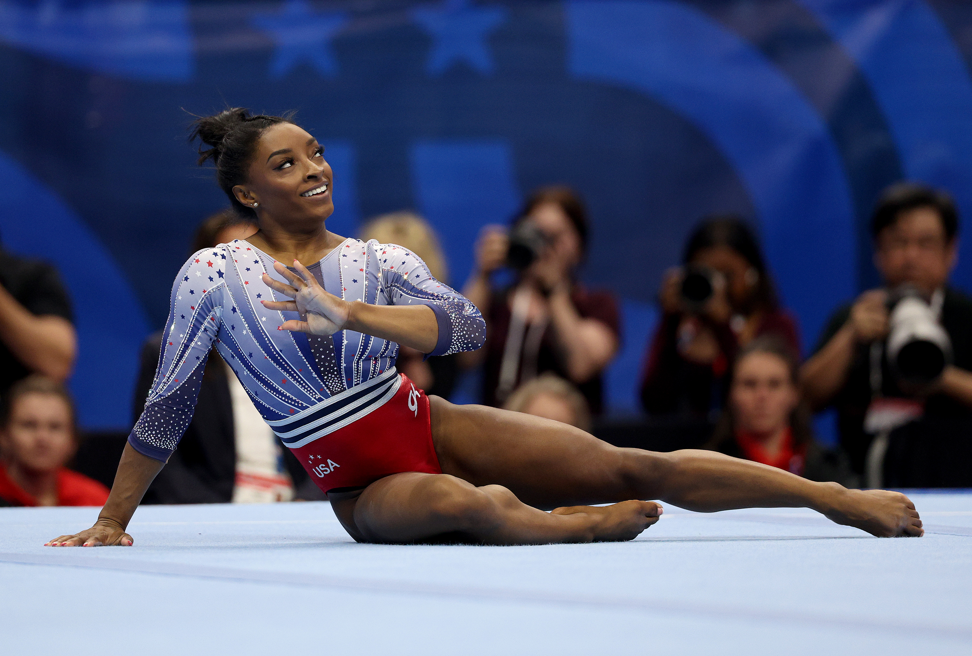 Simone Biles performs a gymnastics routine on the floor, wearing a leotard. Spectators and photographers are in the background