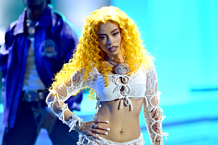 A person performs on stage wearing a lace crop top and shorts. A figure in the background is partially visible. Names are not provided
