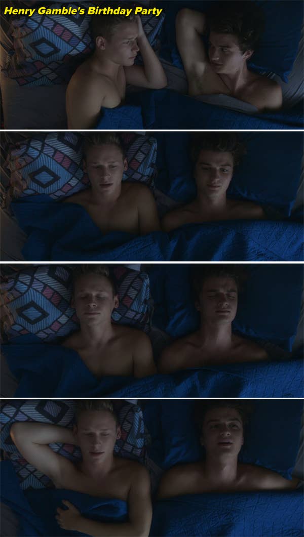 Two shirtless men, one blonde and one brunette, lie together in bed, facing each other and then lying on their backs, sharing a quiet and intimate moment