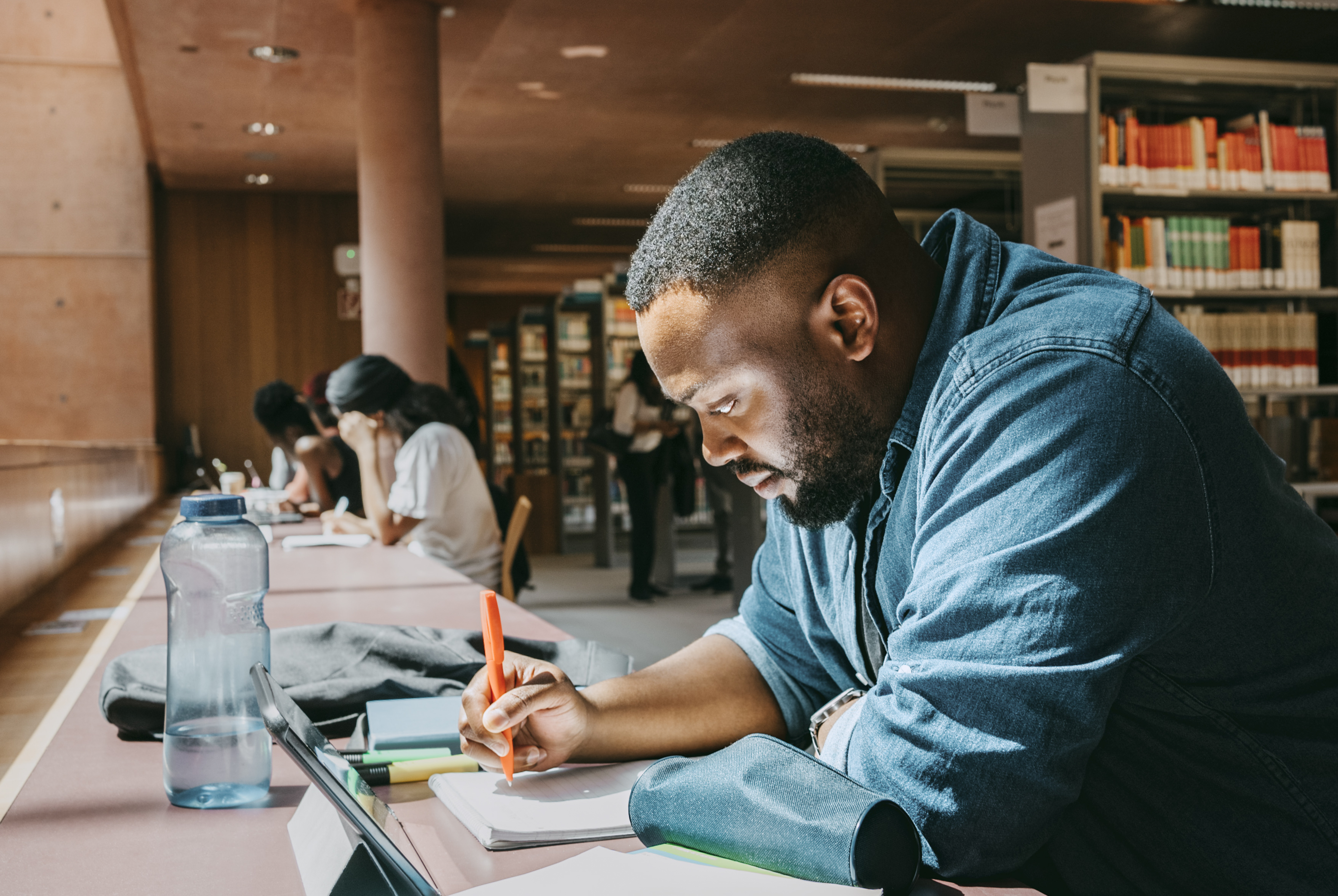 A man is sitting at a desk in a library, focusing on writing in a notebook with a pen. Other people are also studying in the background