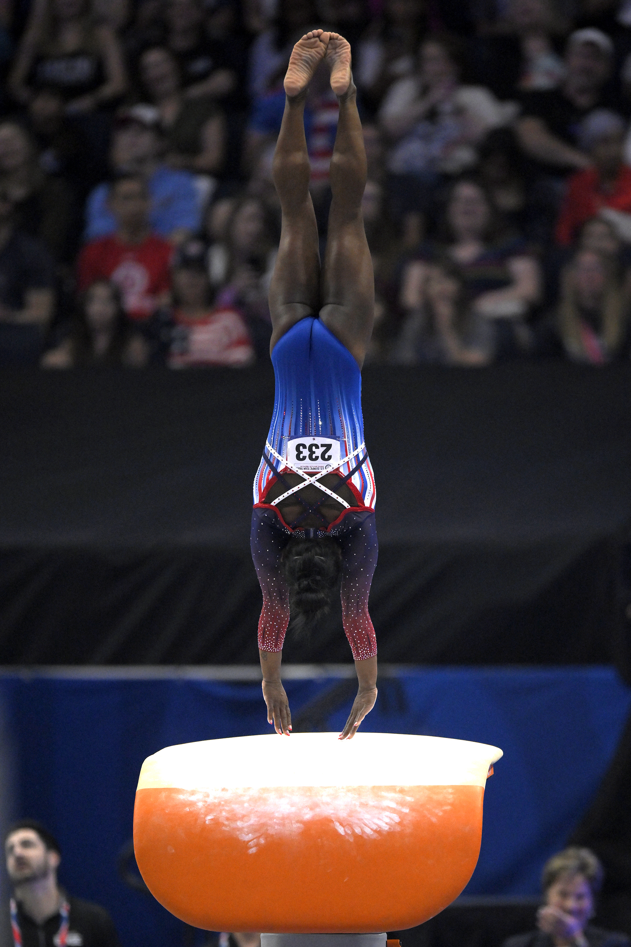 Simone Biles performs a handstand on a gymnastics vault during a competition, wearing a red, white, and blue leotard with the number 233