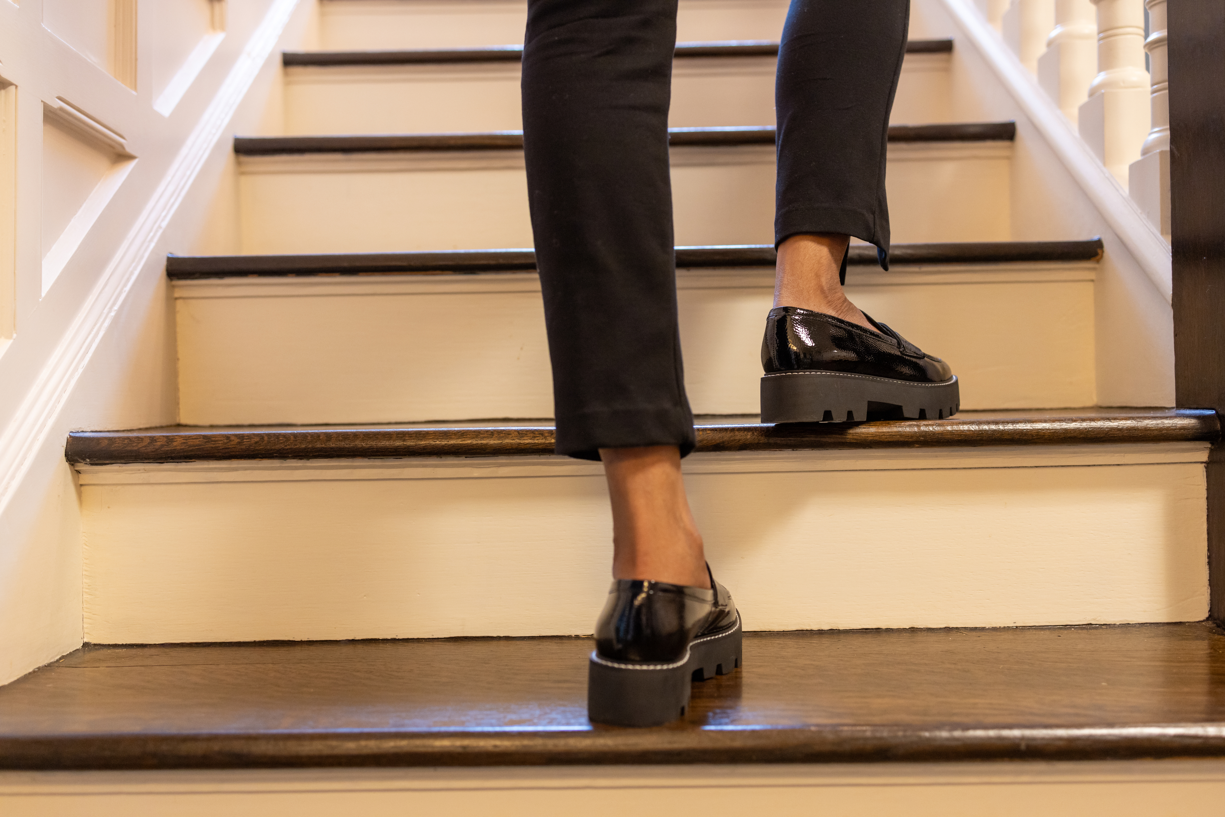 A person wearing black pants and patent leather loafers walks up a wooden staircase. The image focuses on the lower legs and shoes