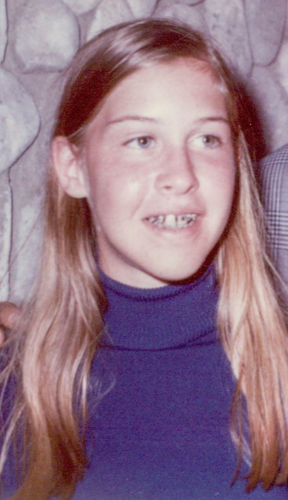 A young girl with long blonde hair and wearing a turtleneck sweater smiles, showing her braces