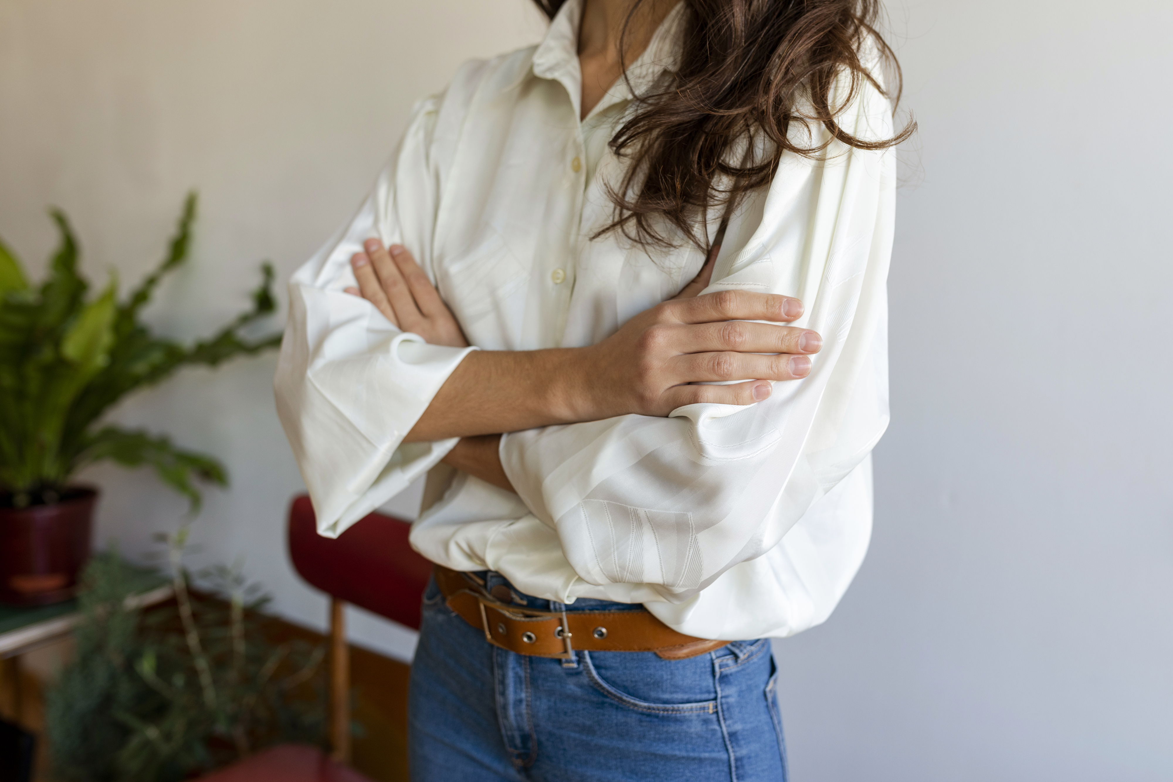 Person with long hair, wearing a light blouse and blue jeans, stands with arms crossed in a casual setting