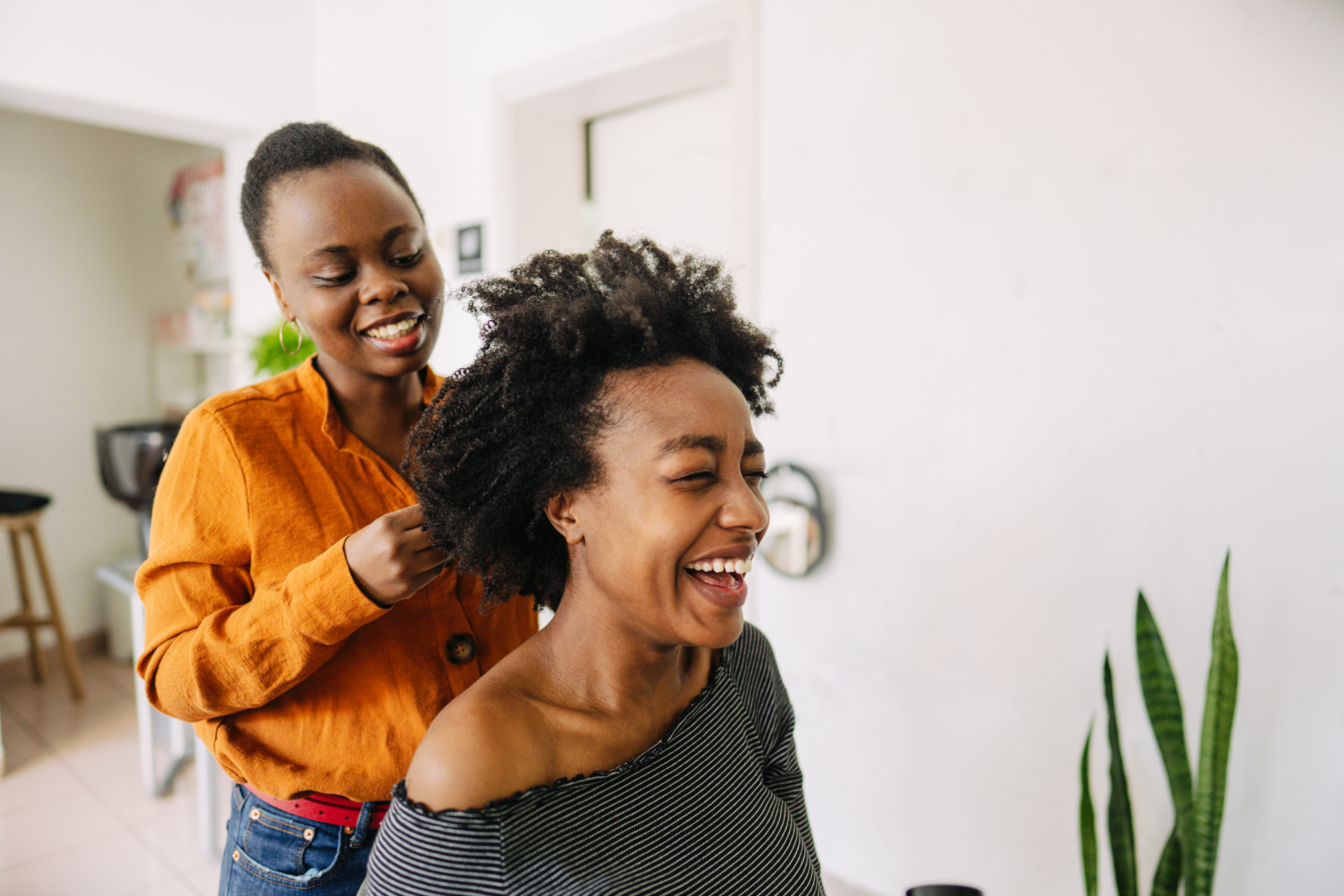 Two women are in a home setting, smiling and laughing as one styles the other's hair. They appear to be enjoying a joyful moment together