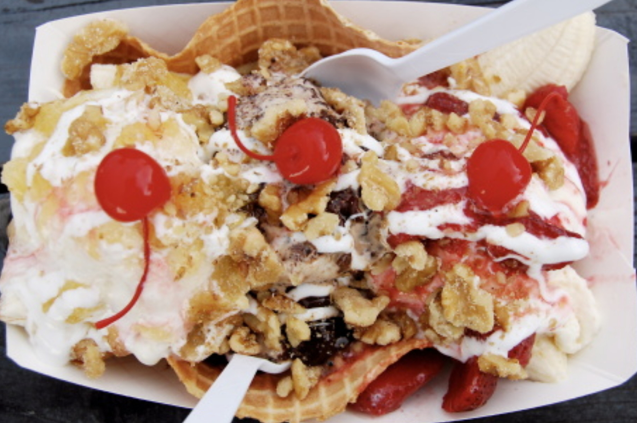 A close-up of a banana split dessert in a waffle bowl, topped with three cherries, crushed nuts, whipped cream, and a variety of ice cream scoops