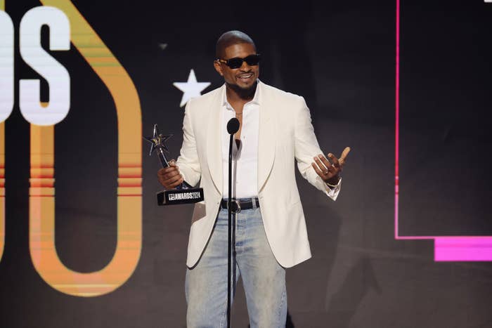 Usher stands on stage holding an award, wearing sunglasses, a white blazer, a white shirt, and jeans