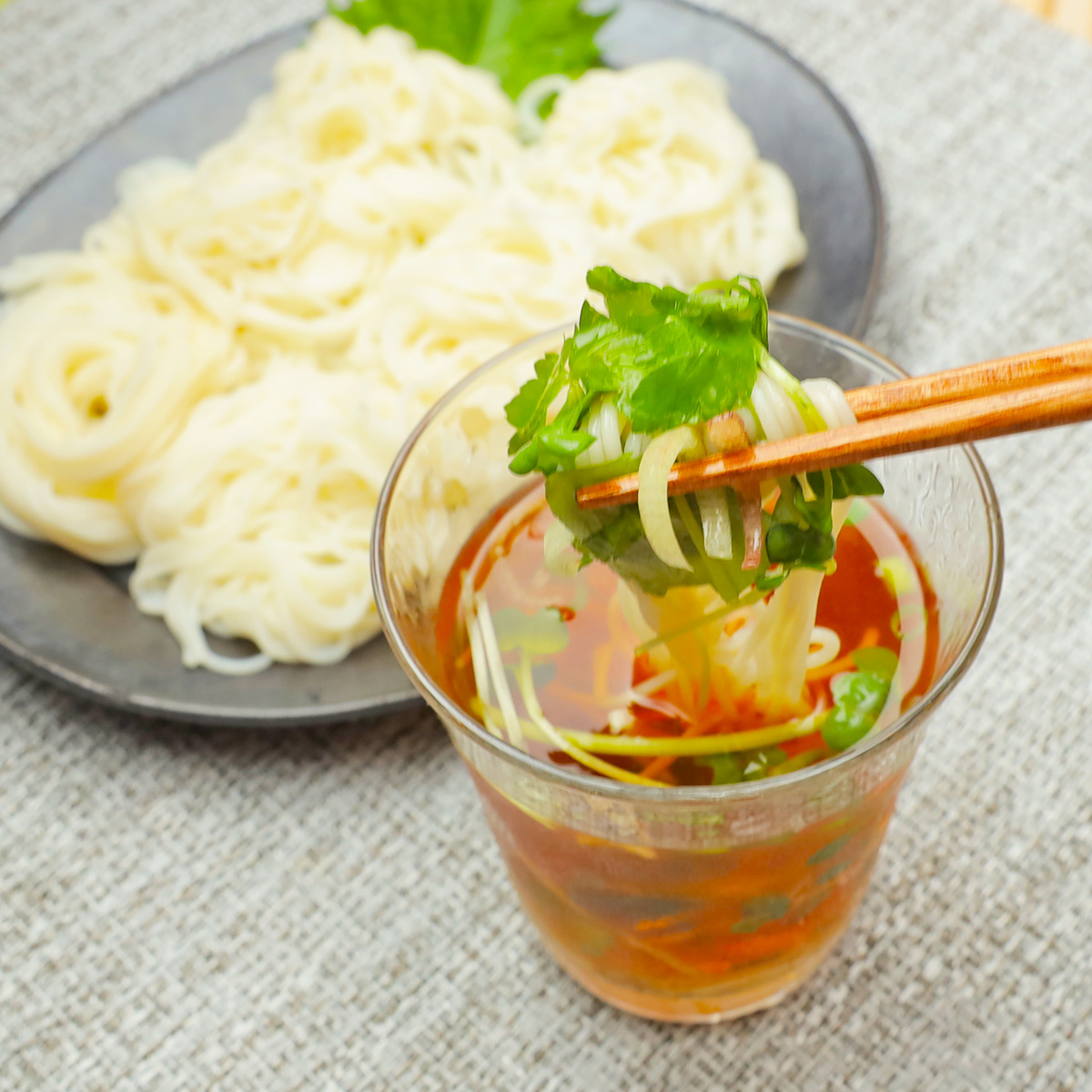 ガラスのカップに入った麺を箸で持ち上げている様子。バックにお皿に盛られた麺が見えます。