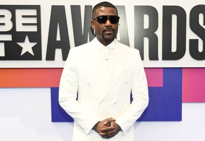 Ray J in a white suit with black sunglasses at the BET Awards, posing in front of a sign with &quot;BET AWARDS&quot; text