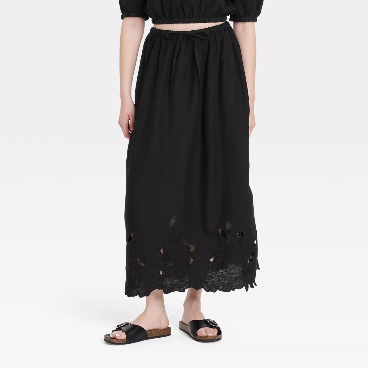 Person wearing a black maxi skirt with a floral cutout hem and black sandals, standing against a plain background. Face is not visible