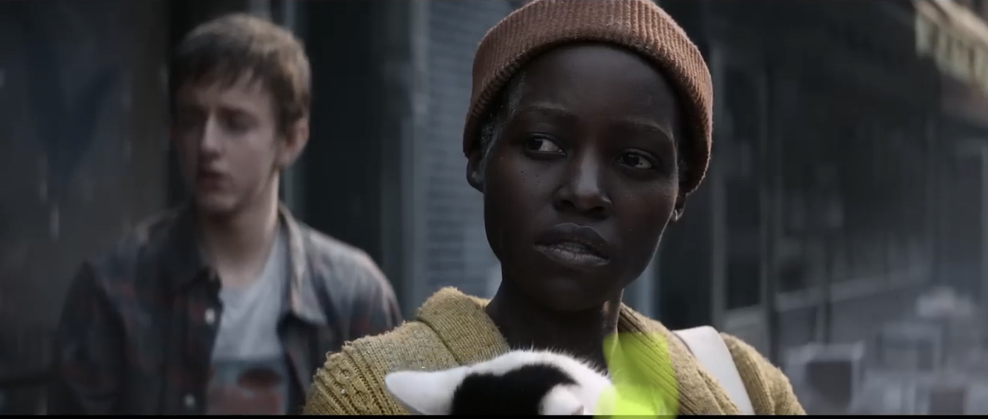 Danai Gurira in a yellow cardigan holding a cat, with an out-of-focus person in the background