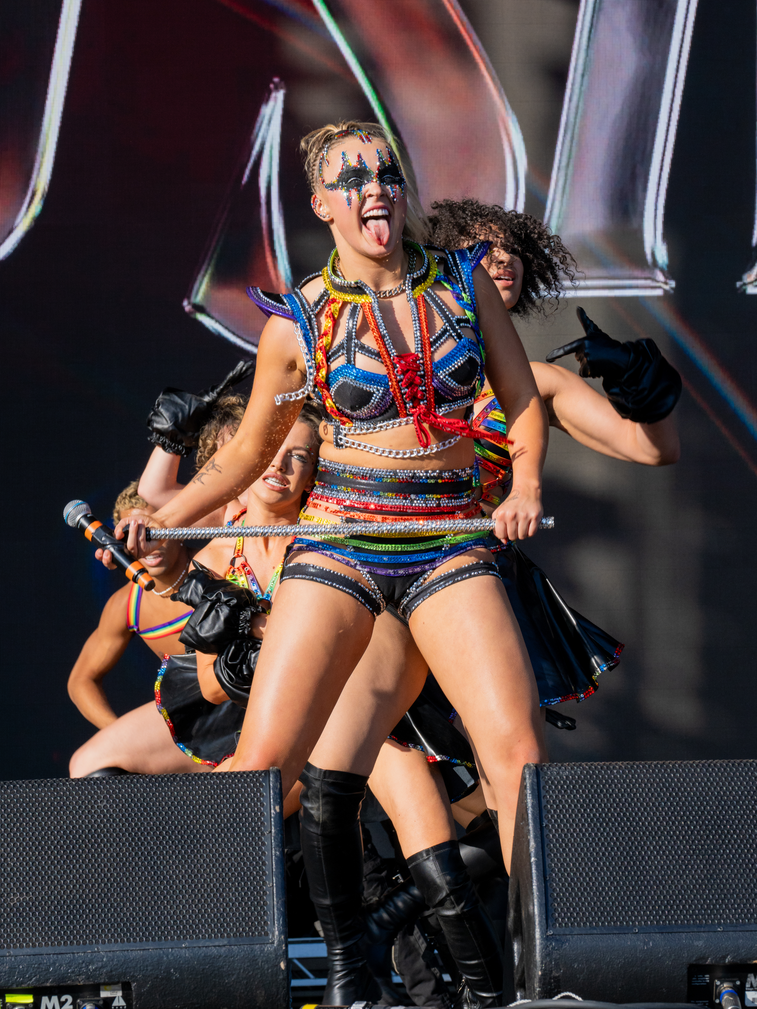 JoJo Siwa performing enthusiastically on stage, wearing a multicolored outfit with beaded fringe, surrounded by backup dancers in coordinating costumes