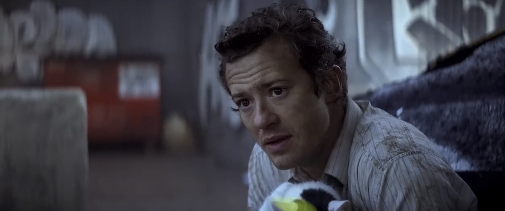 A man wearing a white-striped shirt stares intently while sitting in a dimly lit area with graffiti on the walls in the background