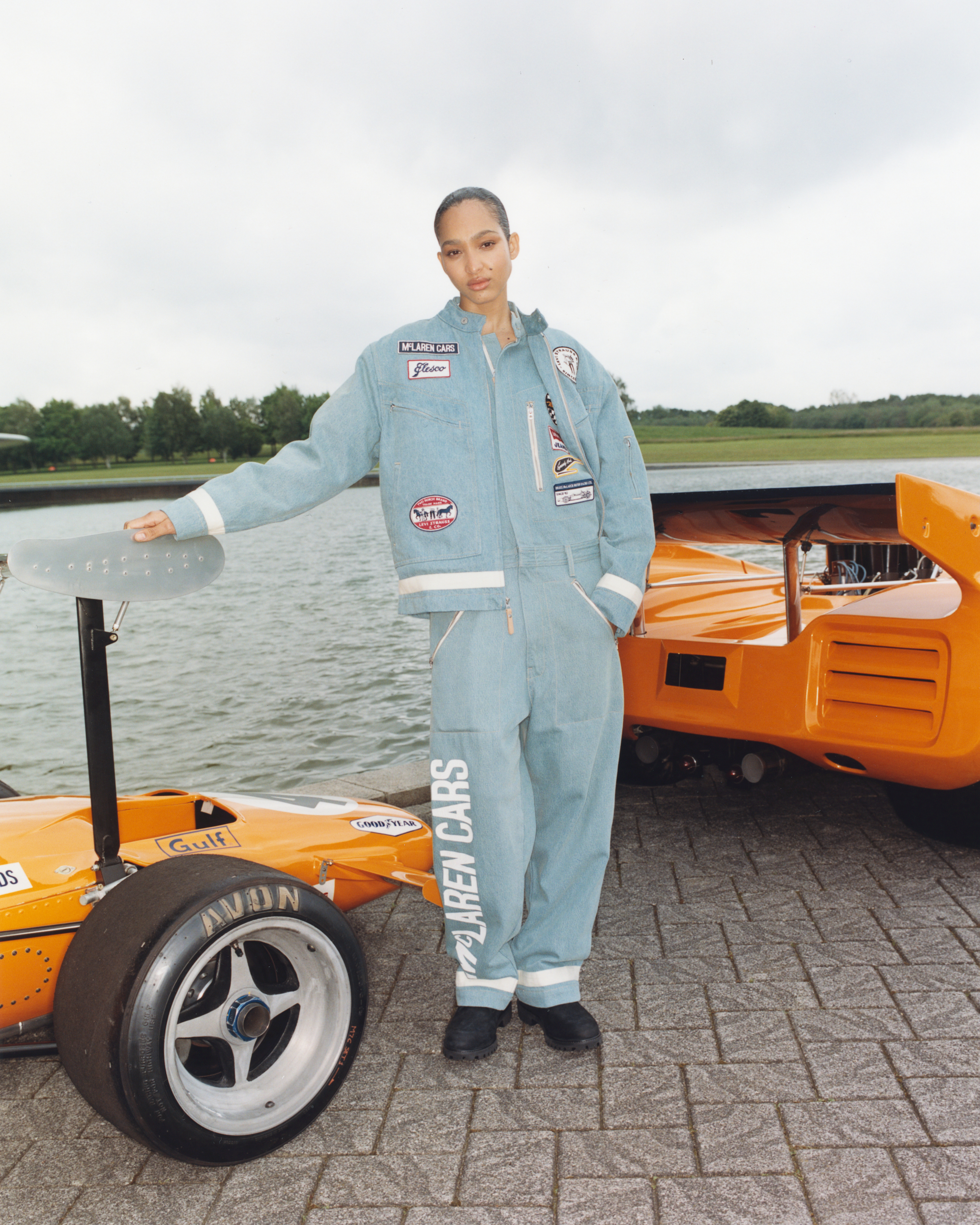Model in a denim jumpsuit stands beside an orange race car near water. The jumpsuit is adorned with various patches. Trees are visible in the background