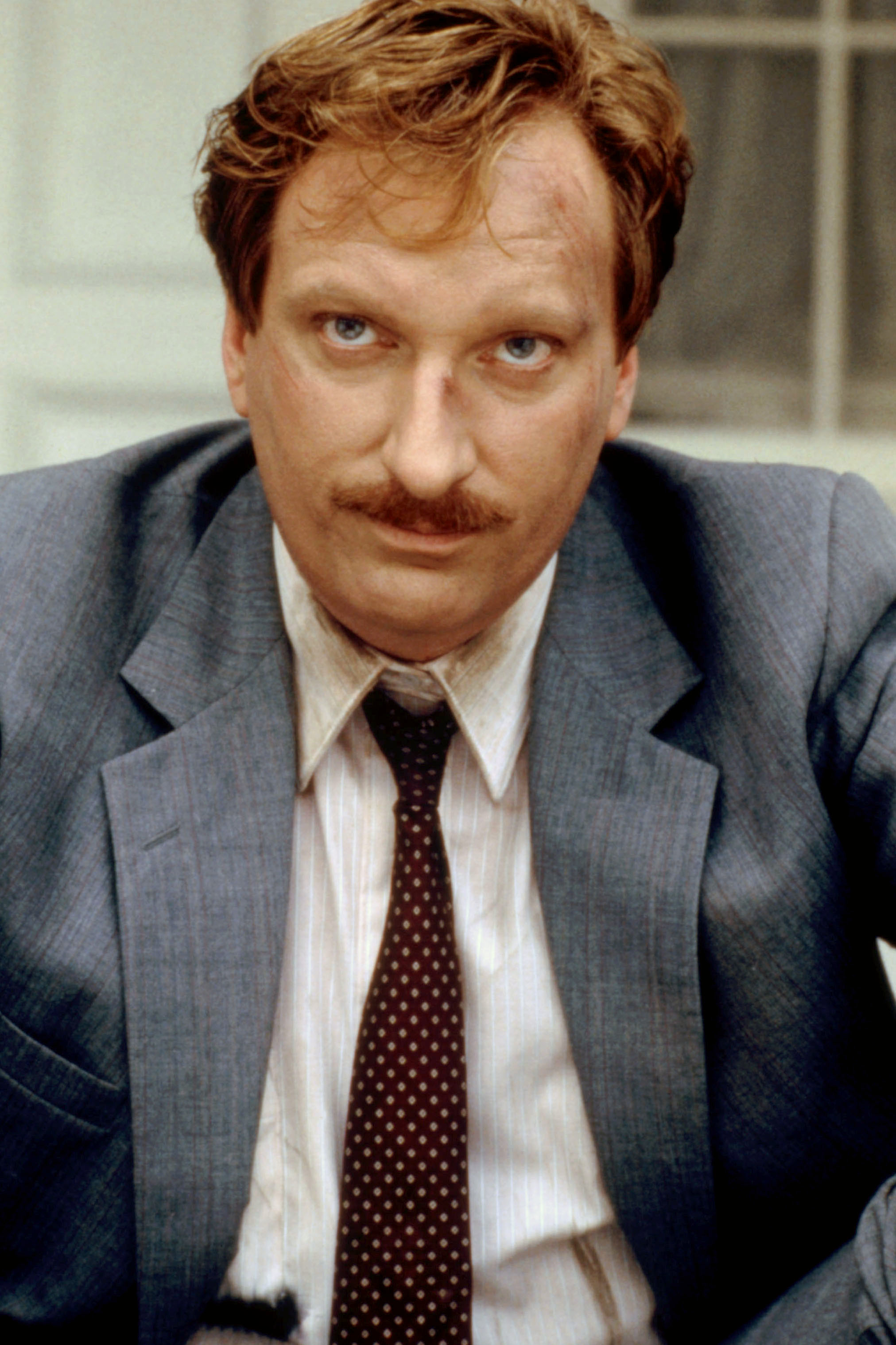 A man with a mustache in a gray suit jacket, white shirt, and dark polka dot tie looks intensely at the camera