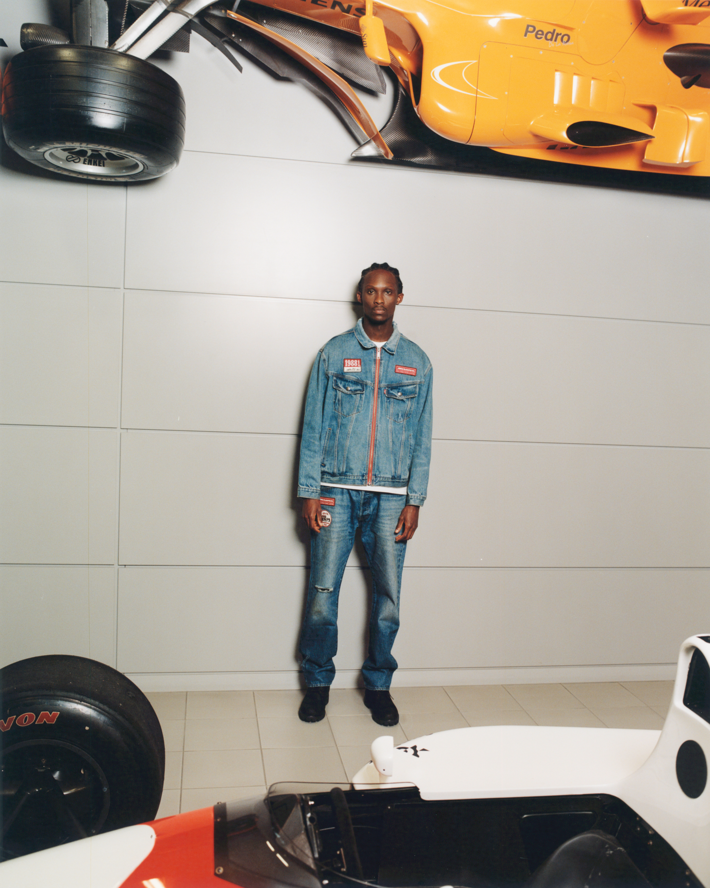 Person in denim jacket and jeans stands in front of a wall with racing car parts displayed