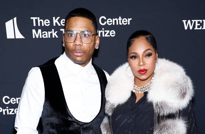 Nelly and Ashanti pose on the red carpet at an event. Nelly wears a white shirt with a black vest, while Ashanti is in a fur-trimmed coat and black dress
