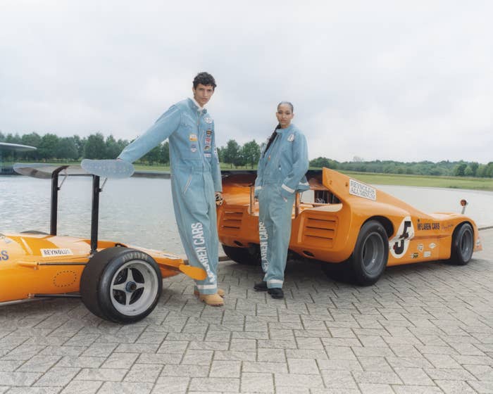 Two models stand in front of racing cars wearing full-body denim jumpsuits with patches, posing near a scenic body of water
