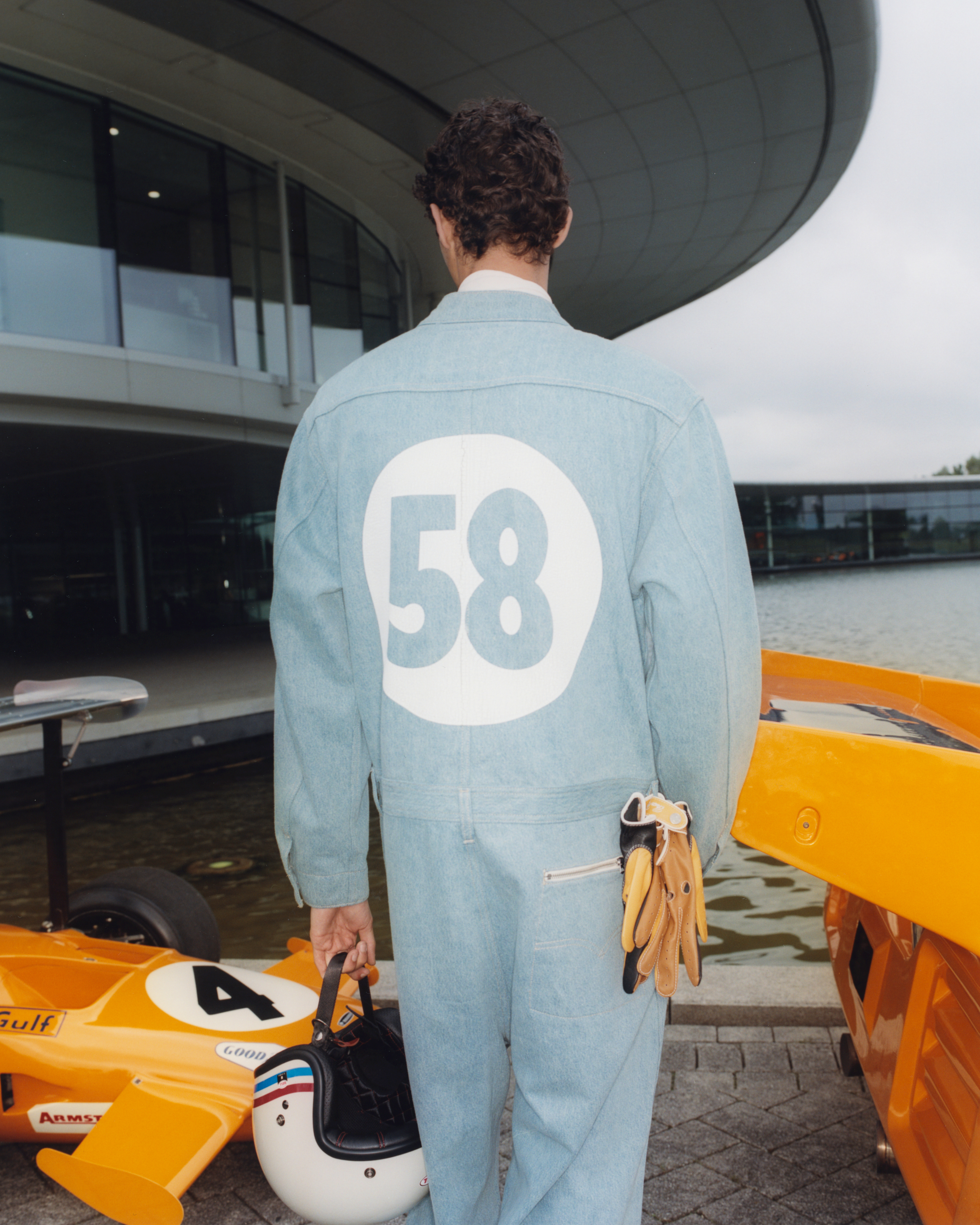 A person is standing with their back to the camera wearing a denim jumpsuit with the number 58 on it, holding gloves and a helmet next to an orange car