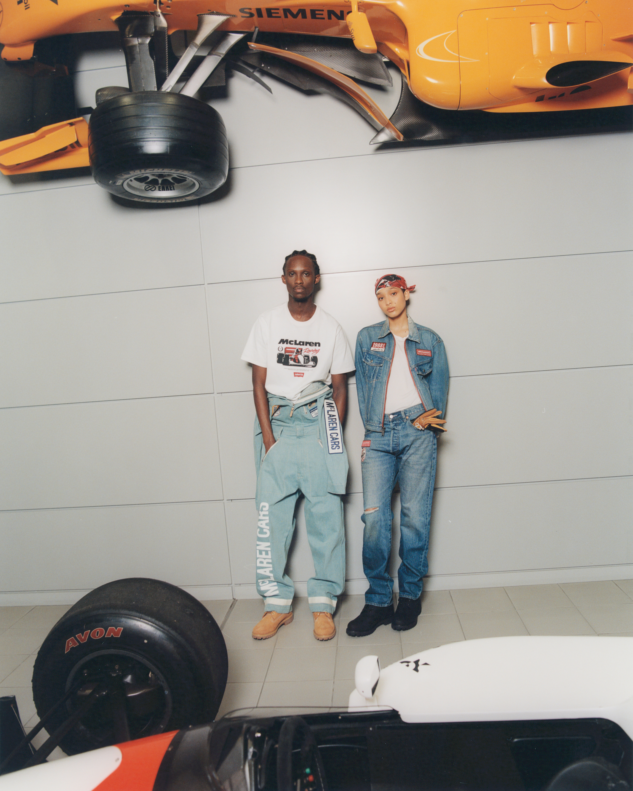 Two people standing in front of cars: one in a printed t-shirt and overalls, the other in a denim jacket and jeans with a headscarf