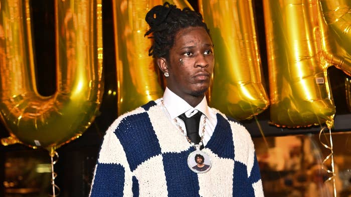 Young Thug wearing a blue and white checkered sweater, standing in front of large gold balloons at an event