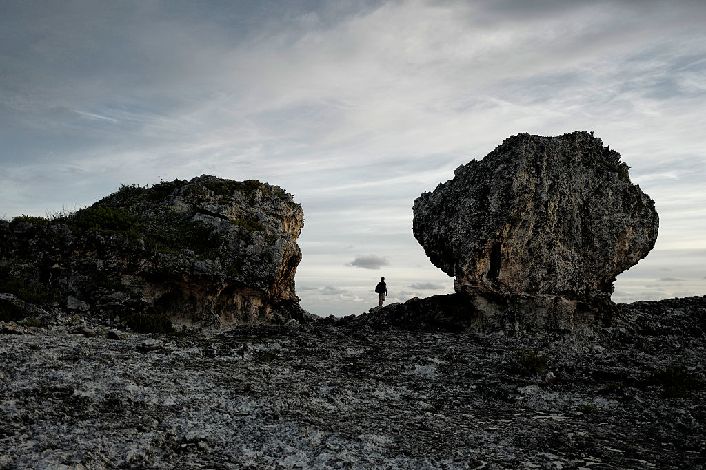 A person stands between two large rock formations on a rugged landscape, silhouetted against a cloudy sky