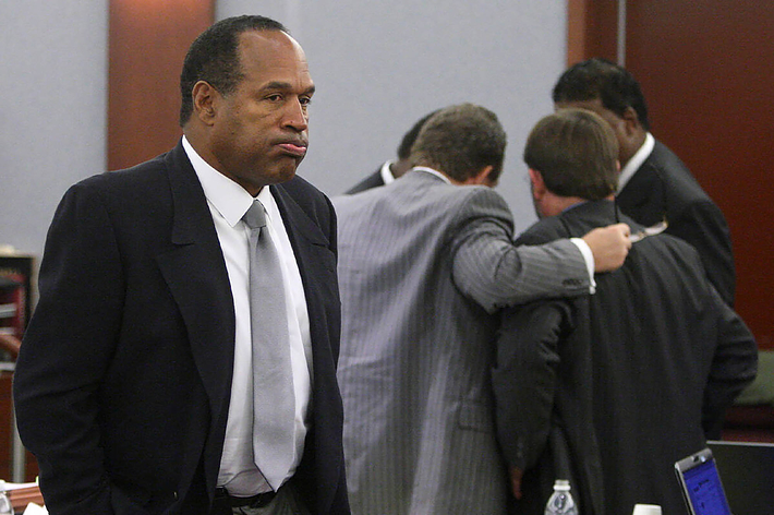 O.J. Simpson stands in a courtroom wearing a dark suit and white shirt, while three men in suits huddle in the background