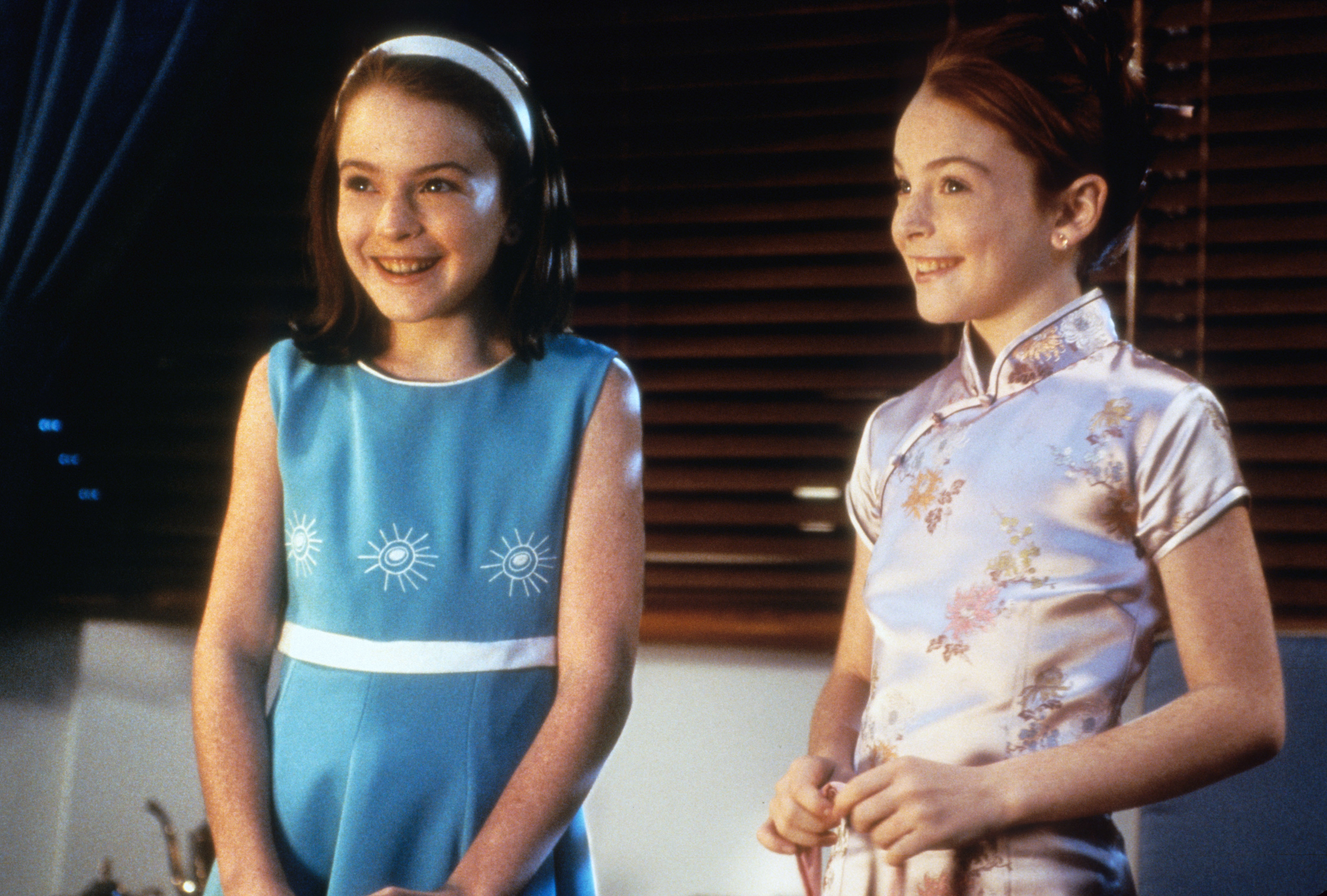 Lindsay Lohan, in a blue dress with white sun patterns, stands next to Lindsay Lohan, in a floral cheongsam, both smiling in a scene from "The Parent Trap"