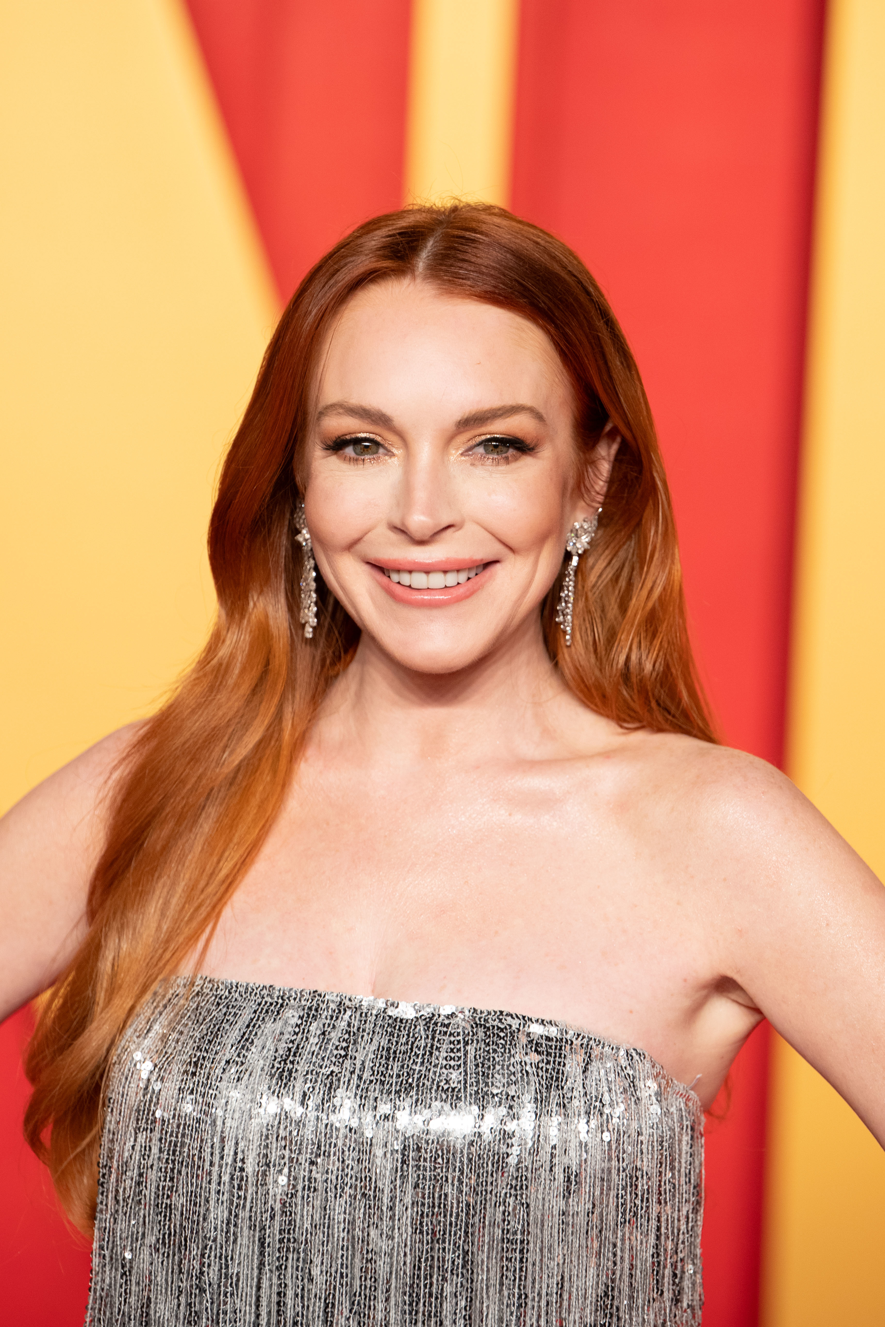 Lindsay Lohan smiling on the red carpet, wearing a strapless dress with fringe and diamond earrings