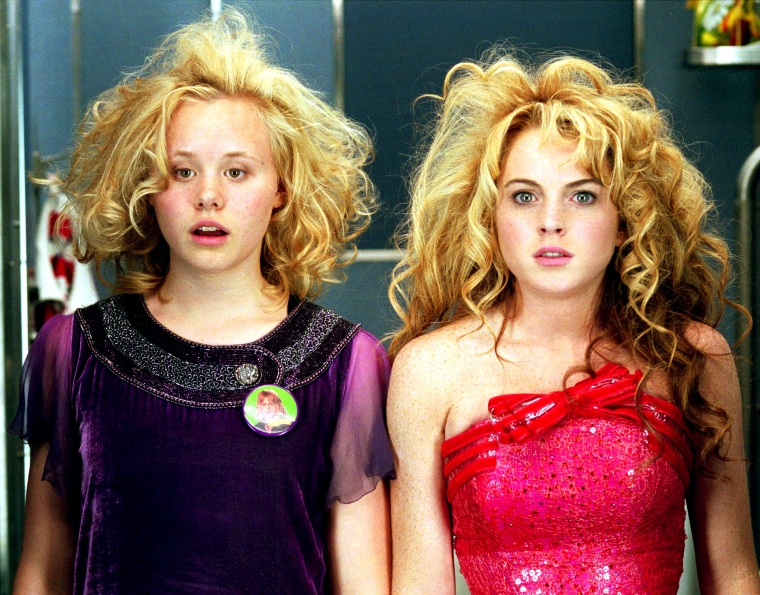 Alison Pill and Lindsay Lohan with tousled hair, standing together. Allison is in a purple dress with a round pin, and Lindsay wears a sparkling strapless pink dress