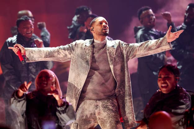 Usher performs on stage in an elaborate suit while backup singers in dark outfits sing enthusiastically behind him