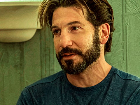 Jon Bernthal with his arms crossed, wearing a textured long-sleeved shirt. He has a beard and medium-length hair, set against an indoor backdrop